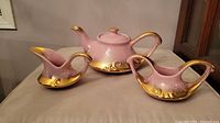 Pink and gold 1960s Aladdin teapot, sugar bowl, and creamer shown side by side on a beige surface.