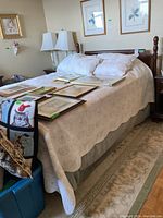 Front and side view of bed frame with headboard showing the bed dressed with beige bedding and decorative pillows along with framed pictures laid out on mattress.