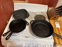 Four cast iron skillets and two metal baking racks arranged on a stove. Skillets vary in size and shape including a square grill skillet.