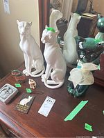 Photo of multiple cat figurines including two large white ceramic cats, small plaques, and smaller figurines on table
