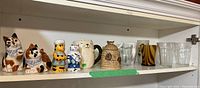Shelf with cat-themed ceramic salt and pepper shakers, creamer and sugar containers, a ceramic beehive honey pot, assorted glass tumblers, and a large cat mug.