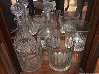 Five glass barware items including two cut glass decanters and three clear glass ice buckets shown against a mirrored surface.