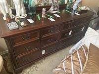 Front view of mahogany dresser with 8 drawers and metal handles, showing wear and scratches on the surface. Various decorative figurines and tags on top.