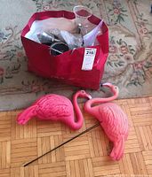 Two pink plastic flamingo lawn ornaments with metal stakes lying on parquet floor near red bag with garden lights.