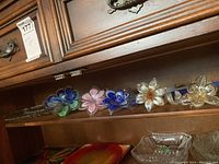Top shelf showing six glass flowers in various colors and a glass platter below.