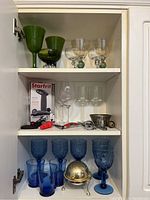 Three white shelves with various glassware and barware items arranged. Mostly green, clear, and blue glassware with bar tools on middle shelf.