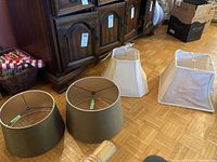 Four fabric lamp shades on wooden floor in front of wooden furniture: two olive green tapered drum shaped shades and two cream square shaped shades with metal frame.