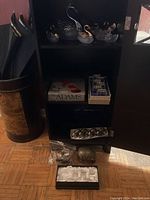 Cabinet with three shelves showing swan figurines on top, boxes and trays with napkin rings on middle and lower shelves, loose napkin rings in plastic bag on floor in front.