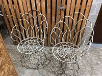 Two white metal decorative patio chairs with scrollwork placed against a wood-slatted wall showing rust and weathering.