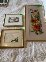Framed needlepoint artwork with vibrant flowers, and two smaller framed floral prints on a quilted white fabric background.