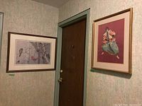 Two framed prints hanging on a hallway wall next to a brown door, showing the positions and appearance of both artworks