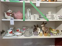 Photo showing ceramic and glass rabbits, cat and pink teapots, cups, and decorative items on shelf.