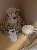 Photo showing stacked plates and teacups with intricate red, black, and gold border pattern and gold trim, identified as Royal Crown Derby.