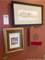 Two framed prints on wall; top one is Saskatoon cityscape with black frame and white mat, lower one is cat print with gold frame and green matting.