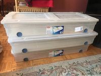 Two stacked translucent plastic underbed storage bins with fitted lids and blue wheels on the side, placed on a wooden floor.
