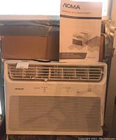 Front view of the white NOMA window air conditioner with box to the left and instruction manual on top.