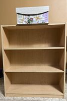 Front view of a rectangular, three-shelf storage unit made of particle board with light wood laminate veneer, empty except for a floral box on top.