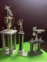 Four trophies shown: large tennis trophy with silver columns, medium tennis trophy with gold figure, table tennis trophy with green and black base, smaller bowling trophy with gold bowler on white marble base.