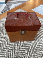Closed vintage textured carrying case with red handle and metal latch, placed on carpet.
