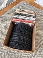 Box filled with about 30 black vinyl 45 RPM records, some in original paper sleeves, arranged upright in a cardboard box on a carpet.