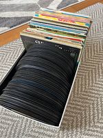 Full box packed with over 100 used 45 RPM records, mostly black vinyl, stacked vertically with sleeves visible at the back
