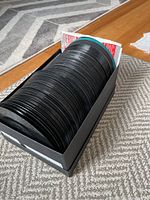 Box containing a large stack of 45 RPM vinyl records vertically placed on a carpeted floor, showing the quantity and condition of the records.