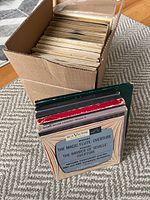 Cardboard box filled with vintage 7 inch record covers and records inside, arranged vertically showing top edges of covers and records.