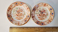 Two ironstone dinner plates with ornate orange and blue border designs and detailed figurative center scenes, shown with a ruler for scale.