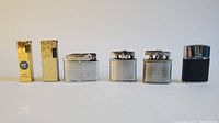 Front view of six vintage pocket lighters in various metal finishes and designs including gold-tone and silver-tone.