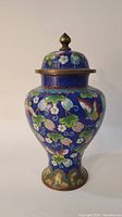 Front and side view of the blue cloisonne jar with lid showing floral and butterfly designs on a blue background. The lid has a metal finial and the base shows remnants of gilt.