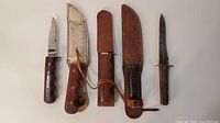 Five cutting tools laid flat with three sheaths visible, varying blade lengths and handle designs