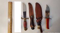 Photo showing four knives with sheaths and handles, ruler for size reference
