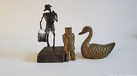 Wide shot showing three metal decorative figures: wire fisherman, brass captain, and brass swan positioned side by side on a white background.