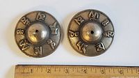 Top view of two round Tibetan cymbal bells with embossed symbols and a hole in the center, placed next to a ruler for size reference.