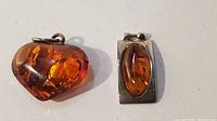 Two amber pendants shown side by side on a white background. One is a polished heart-shaped amber pendant and the other a rectangular silver pendant with an oval amber inset.