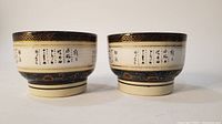 Front view of two Japanese footed ceramic cups side by side showing handpainted characters and gilt trim.