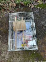 White steel bird cage placed on ground with bagged accessories and wooden perches inside.