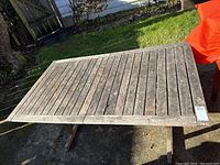Full top view of rectangular wooden outdoor deck table on concrete patio with grass and fence background.