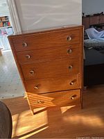 Front view of wood dresser showing five drawers and metal knobs/handles, medium brown finish, standing on hardwood floor.
