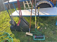 Photo showing two snow shovels, black leaf rake (Ace brand), broom, garden hose, garden cultivator, push lawn sprinkler, and nitrogen probe in an outdoor lawn setting.