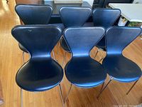 Photo showing 6 black leather dining chairs with chrome legs arranged together on wood floor.
