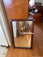 Wood framed mirror leaning against a wood-paneled wall in a room with wood floor and other wooden furniture visible in the background.