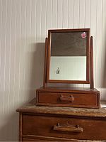 Front view of wooden tabletop mirror with rectangular frame, single drawer, and wooden handle on top of a wooden dresser.