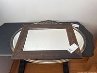 Rectangular mirror with dark wooden frame positioned on top of the oval silver framed mirror on a black table against a white wall.