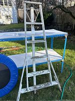Full view of white wooden A-frame ladder with peeling paint and signs of wear, standing outside on grass near a blue table.