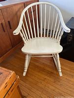 Front view of white wooden rocking chair with spindled back, curved armrests, and contoured seat on hardwood floor near wooden cabinet.