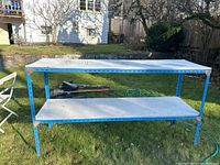Full front view of the two-tier white and blue metal shelf unit set outside on grass with background of house and garden.