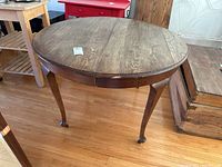 Round oak dining table with three extension leaves, cabriole legs, natural wood finish shown from an angle.