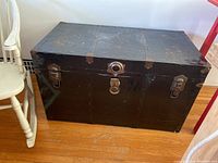 Full view of the black sea chest trunk showing its rectangular shape, riveted metal panels, and metal latches on the front and sides.