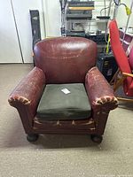 Front view of vintage red leather armchair with olive green cushion showing wear and distressing on leather upholstery and rounded wooden feet.